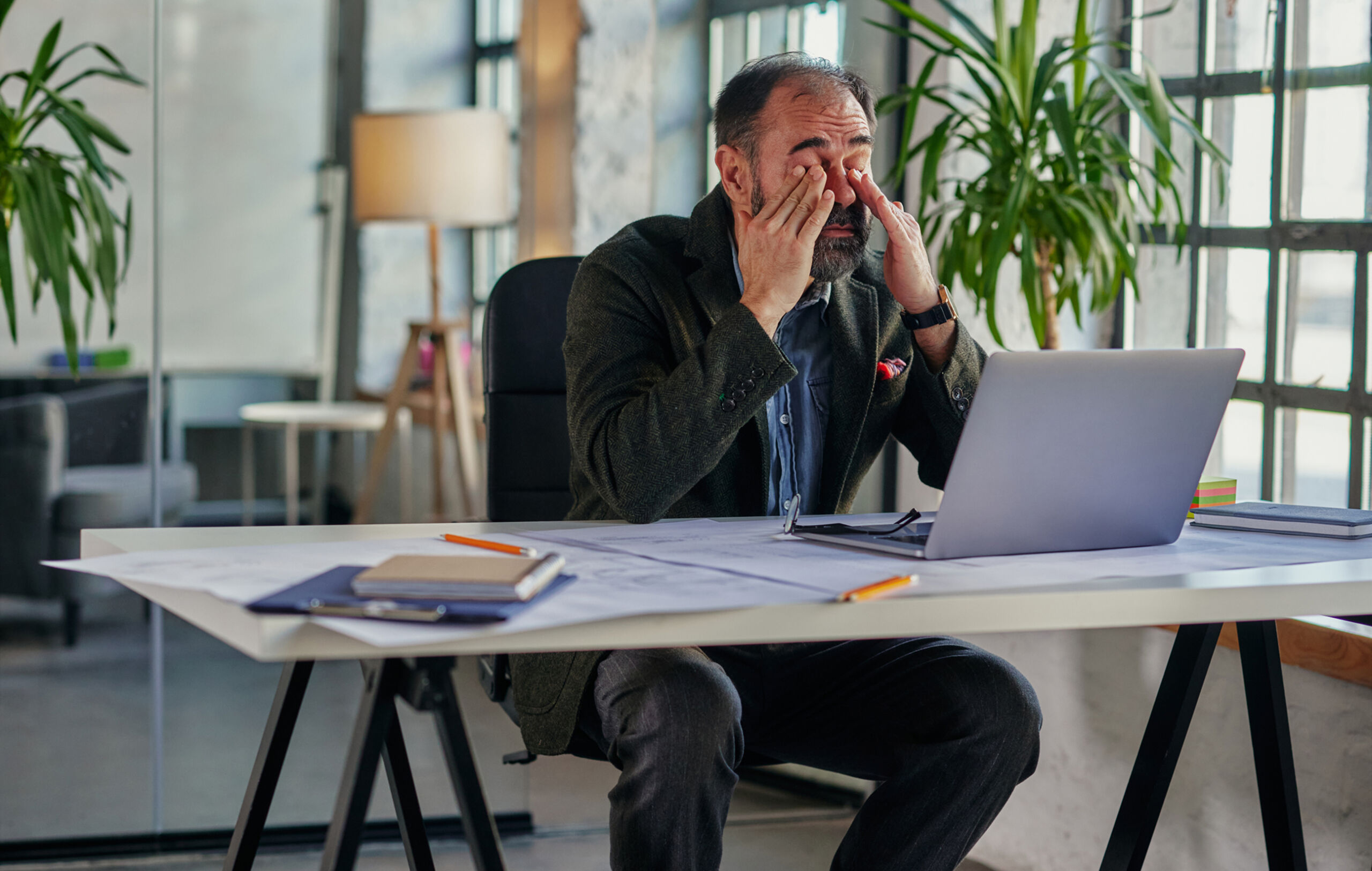 Partner manager rubbing his eyes in modern bright office, staring at computer with green plants around him, worryign because he doesn't know if he can trust his ai with his partners.